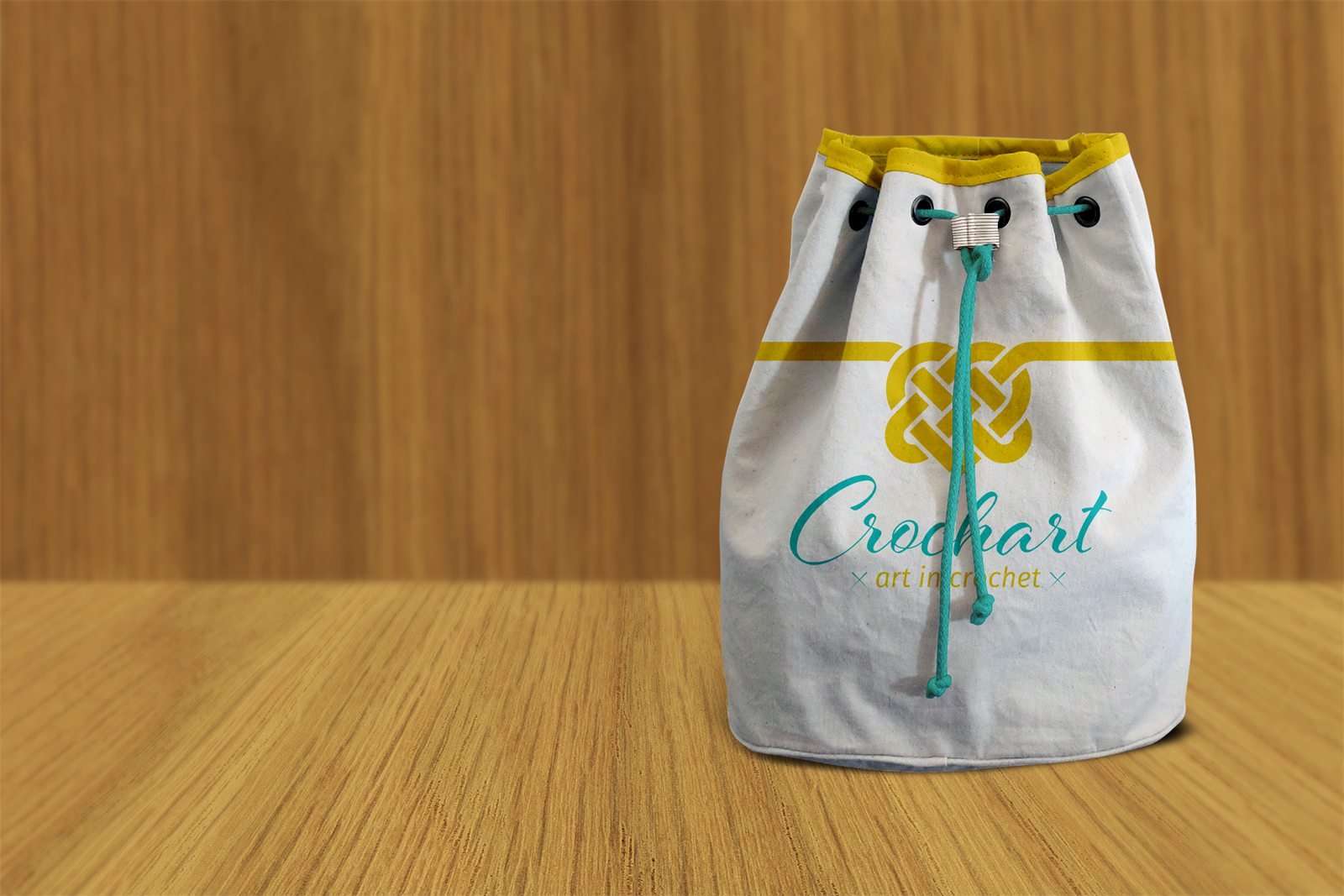 Bolsa-saco de lona crua com borda amarela e cordão verde-água, estampada com o nó e a assinatura da Crochart, sobre uma mesa de madeira