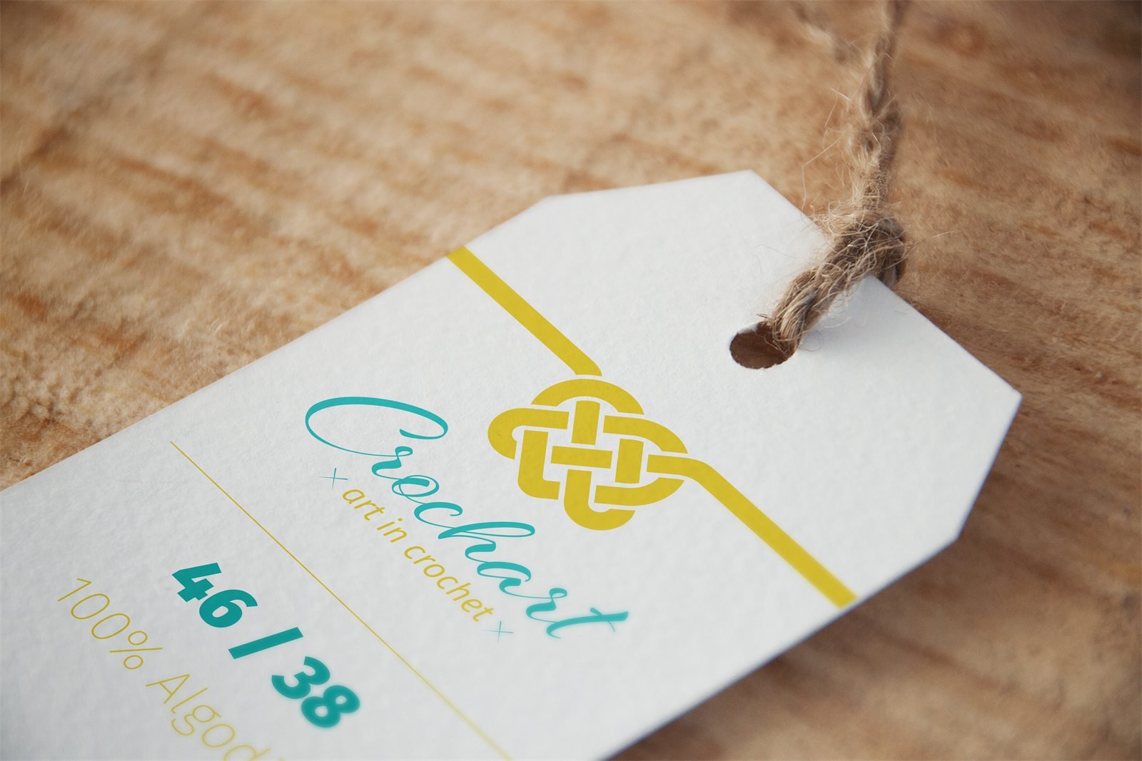 Tag de produto em papel branco, presa por barbante sobre madeira, com a assinatura da Crochart, a numeração 46 / 38 e a composição 100% algodão