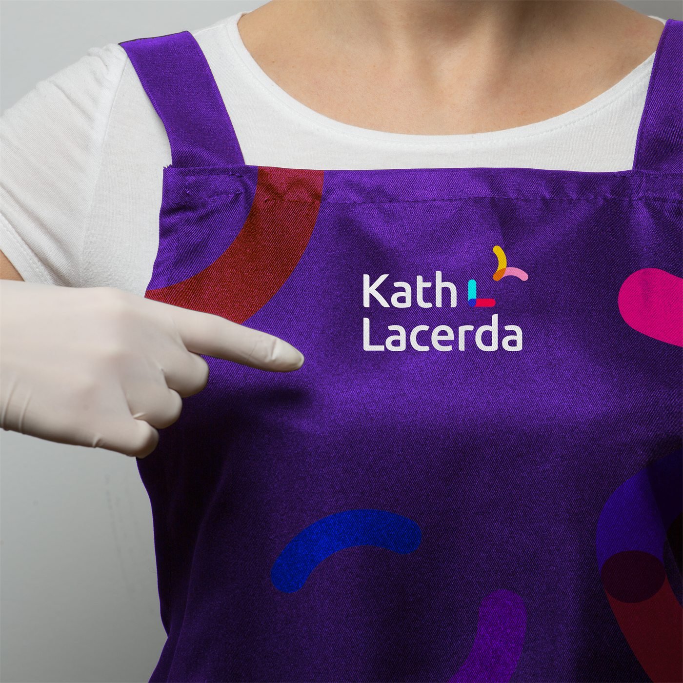Mão de luva aponta a assinatura Kath Lacerda estampada no avental roxo com as formas coloridas da marca