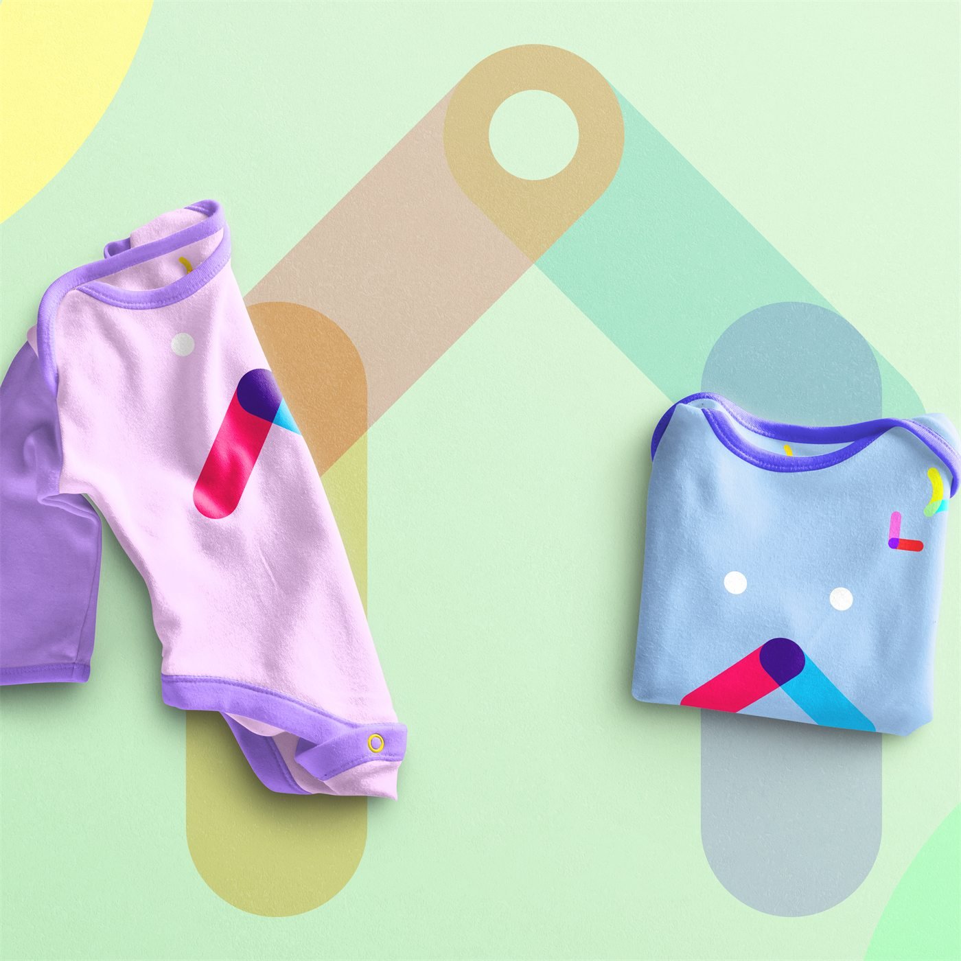 Dois bodies de bebê, lilás e azul-claro, com as formas coloridas da marca, sobre fundo verde-claro