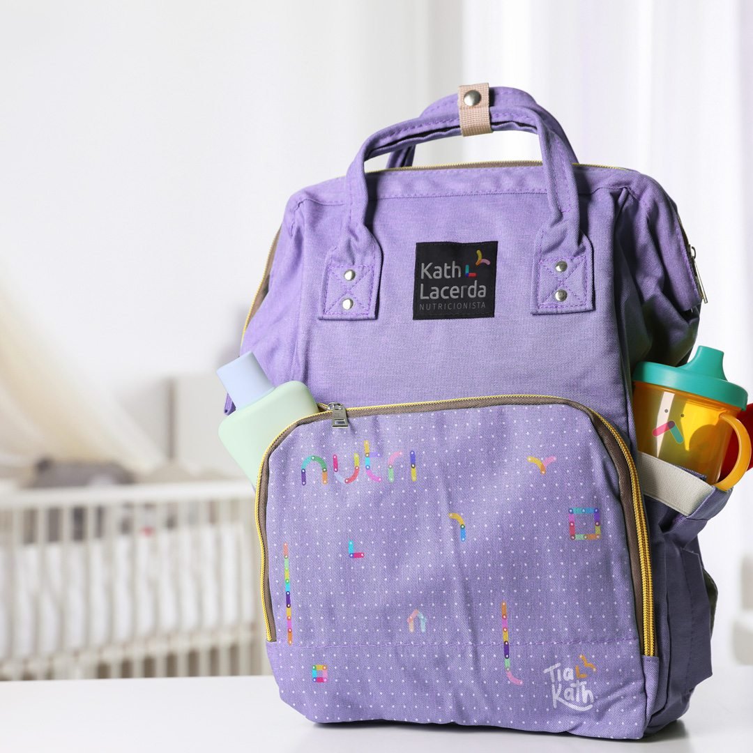 Mochila maternidade lilás com a etiqueta Kath Lacerda e a assinatura Tia Kath, com garrafa e copo nos bolsos laterais