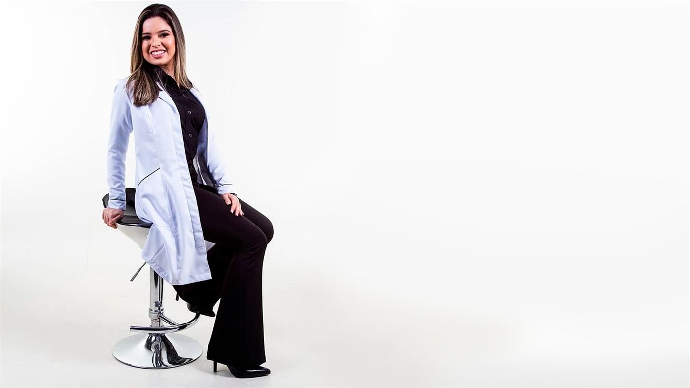 Kathelynne Lacerda, nutricionista, sorri sentada numa banqueta, de jaleco branco sobre roupa preta, em fundo branco