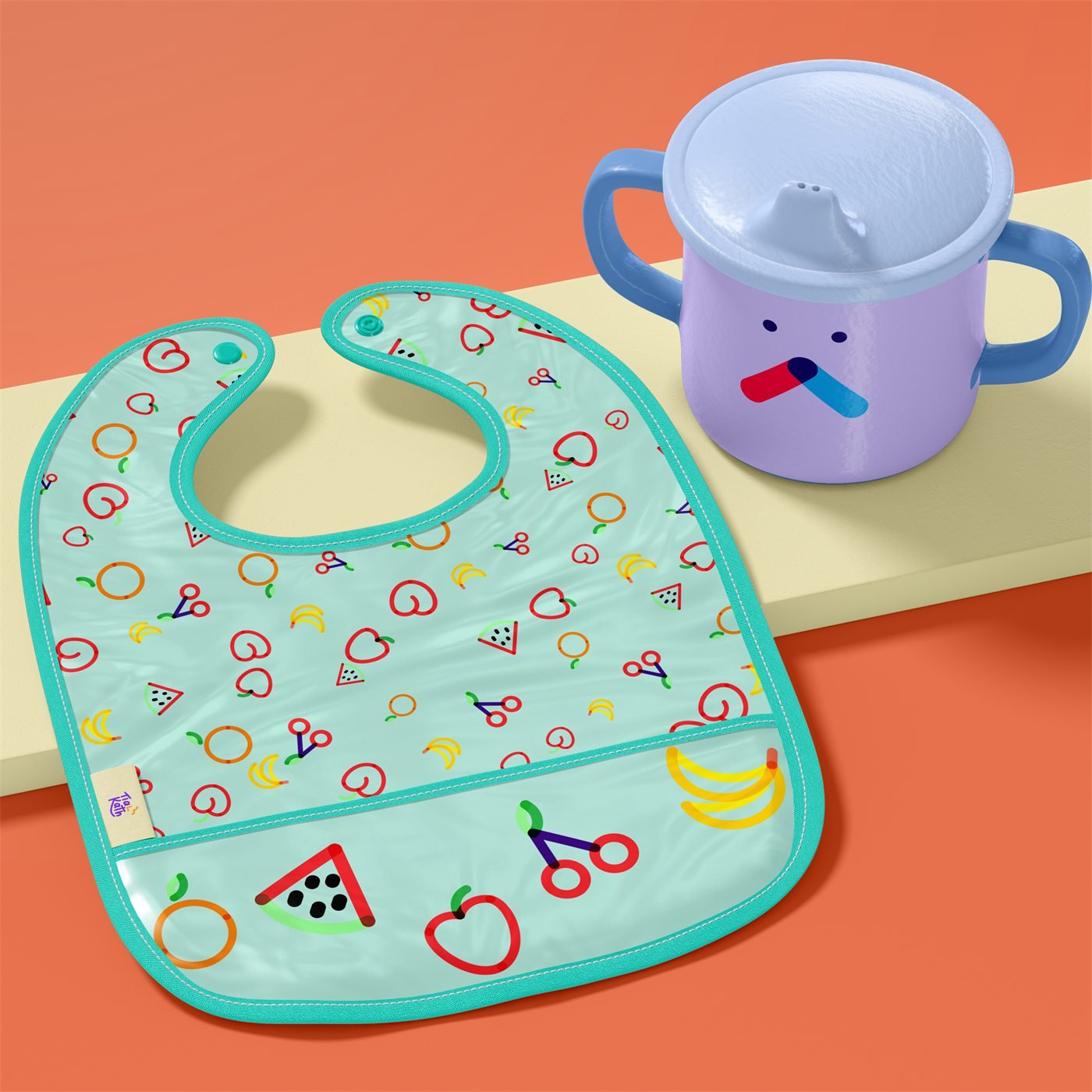 Babador verde-água com estampa de frutas e copo infantil lilás com carinha, sobre fundo coral