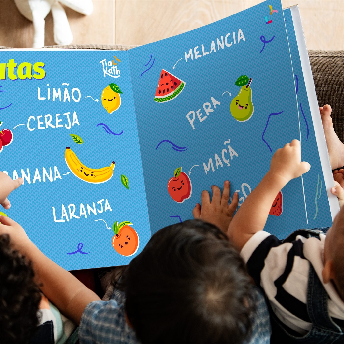 Crianças folheiam o livro de frutas da Tia Kath, com frutas ilustradas e nomeadas: limão, cereja, banana, laranja, melancia, pera e maçã