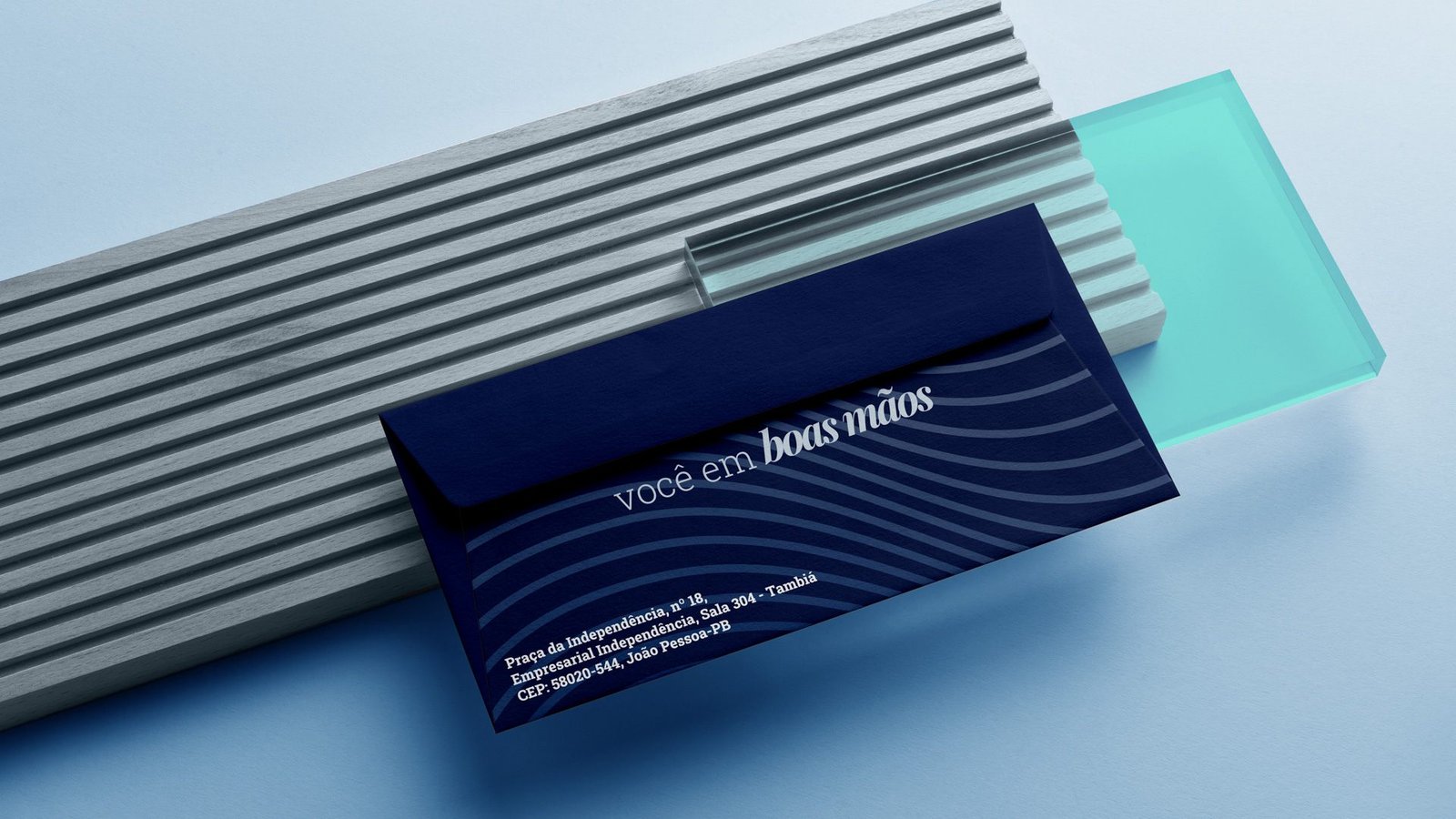Envelope azul-marinho com a frase você em boas mãos e as linhas da marca, apoiado sobre blocos cinza e verde-água