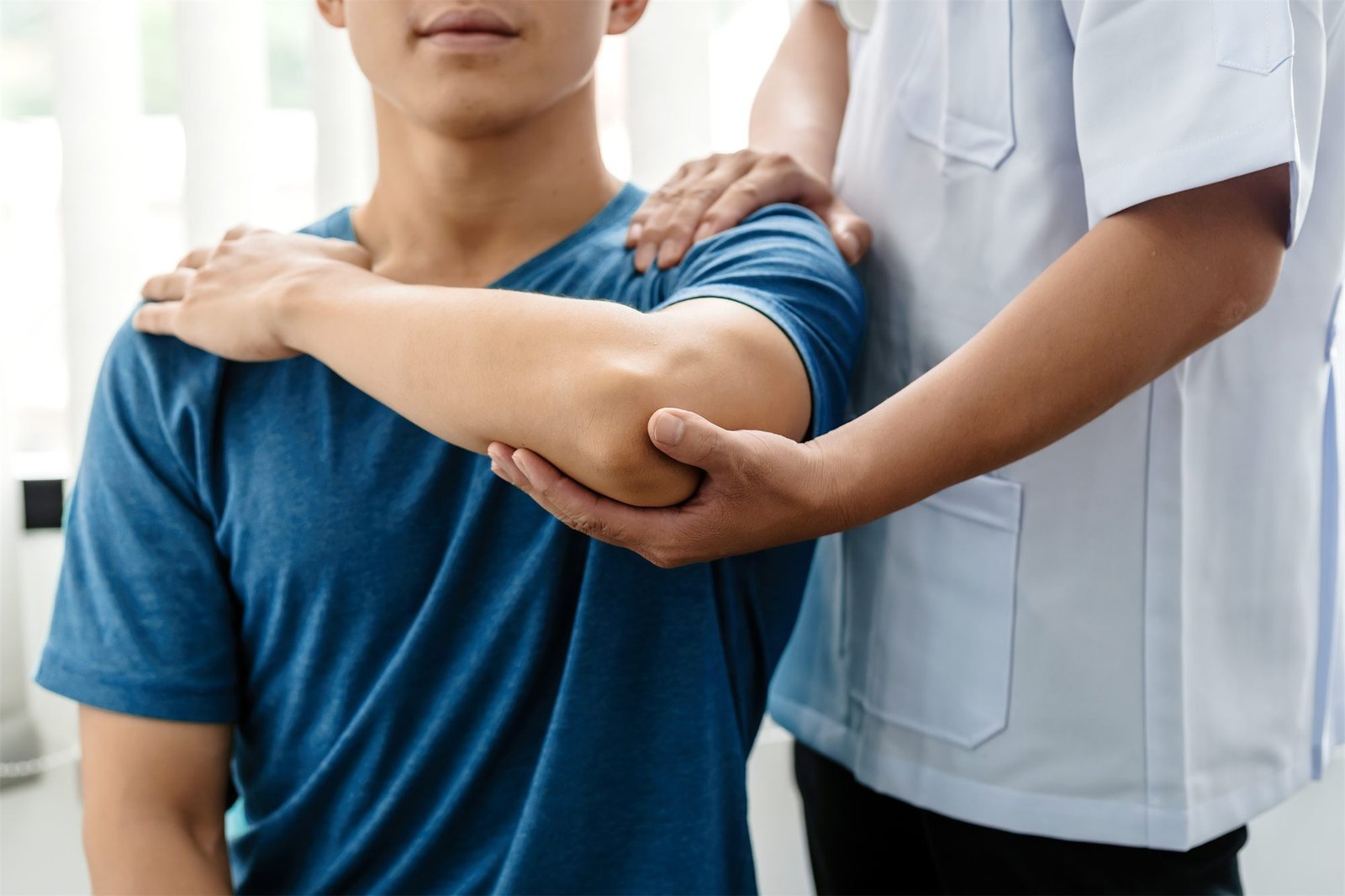 Fisioterapeuta de jaleco apoia o ombro e o cotovelo de um paciente jovem durante um alongamento