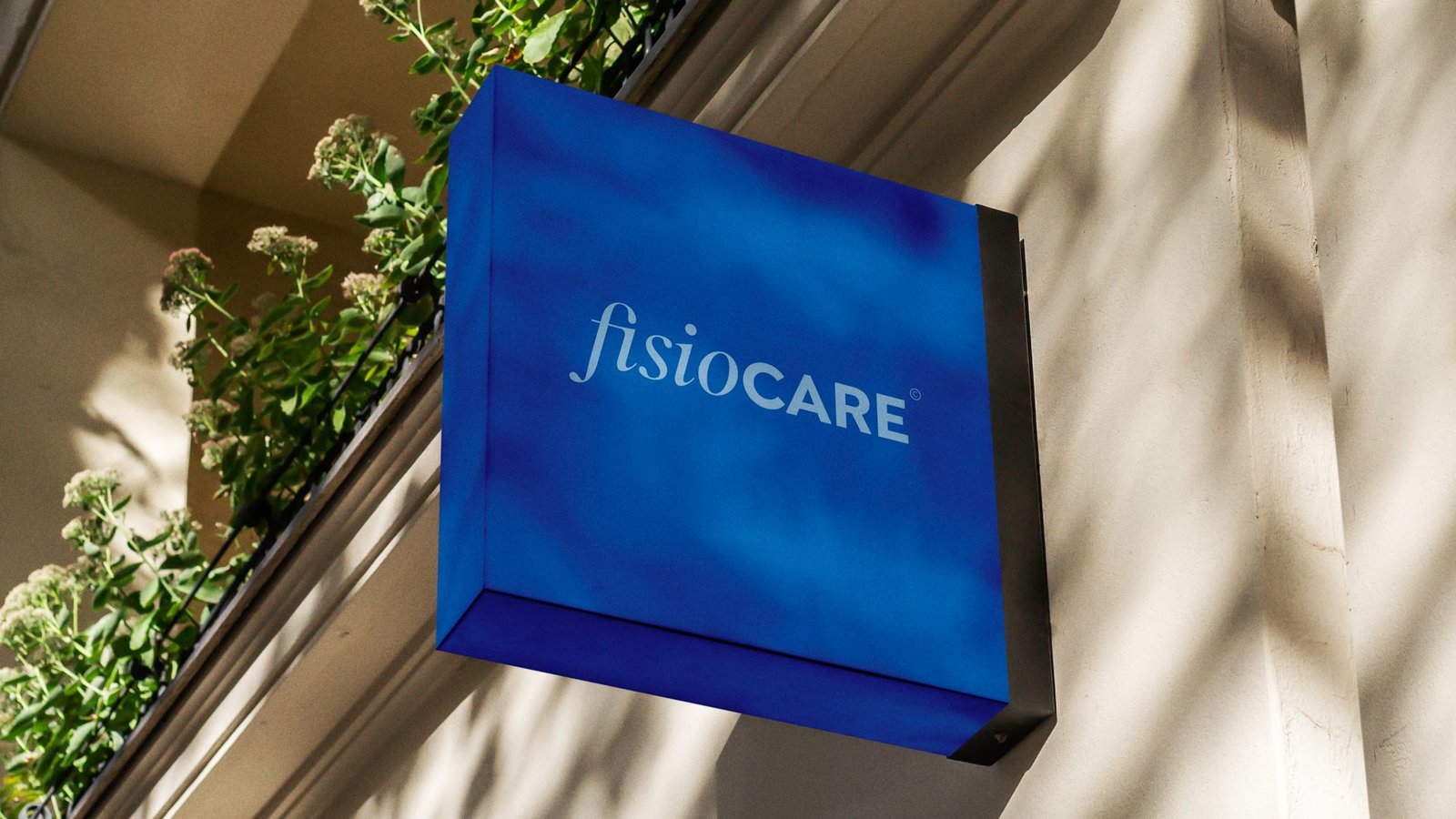 Placa azul com a assinatura fisioCARE presa à fachada, entre plantas e sombras de folhas