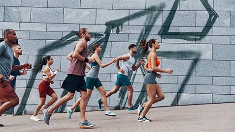 Grupo de pessoas corre junto por uma calçada, diante de um muro com o grafismo da marca